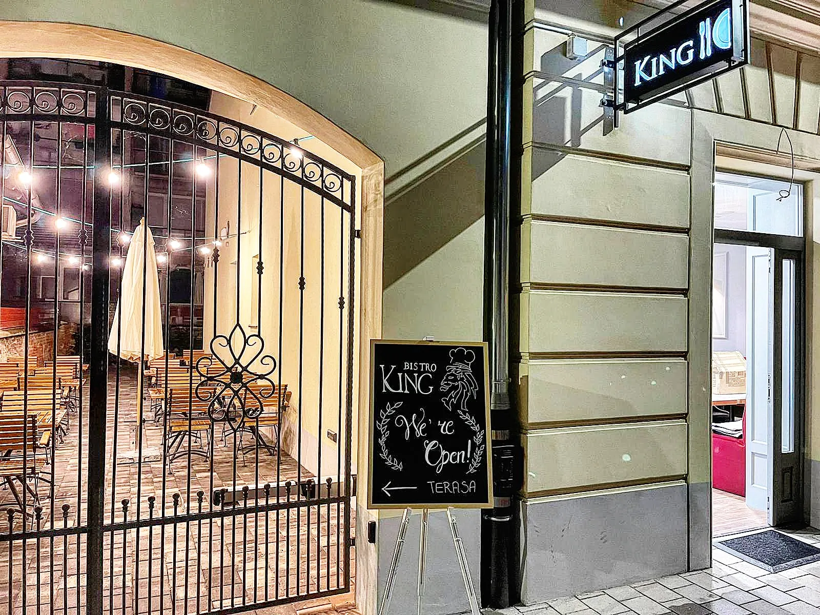 Kapija i ulaz u Bistro King navečer, u dvorištu svijetle lampice, a pored kapije stoji ploča s natpisom Terasa